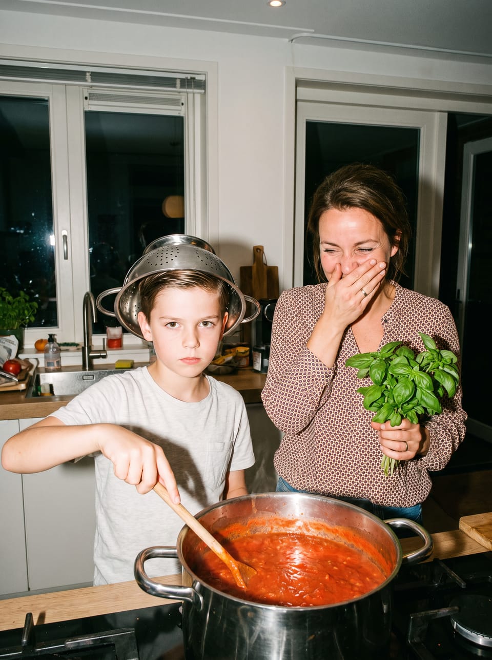 Bram van 9 roert in een pan tomatensaus met een vergiet op zijn hoofd, zijn moeder lacht